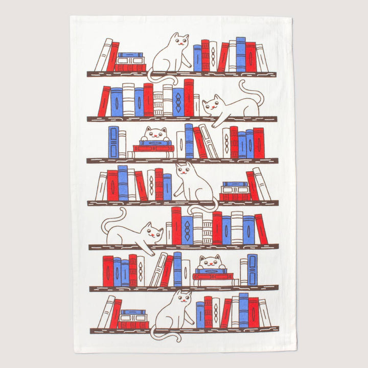 Cotton kitchen towel with cats in various pose on bookcase shelves. Books spines are red and blue. Cates are outlined in dark gray. 