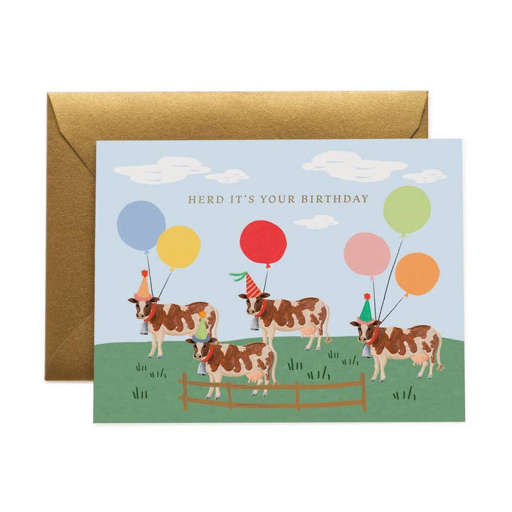 Four cows standing in a field with balloons tied to their necks and wearing party hats under a blue sky with the text 'Herd It's Your Birthday.'