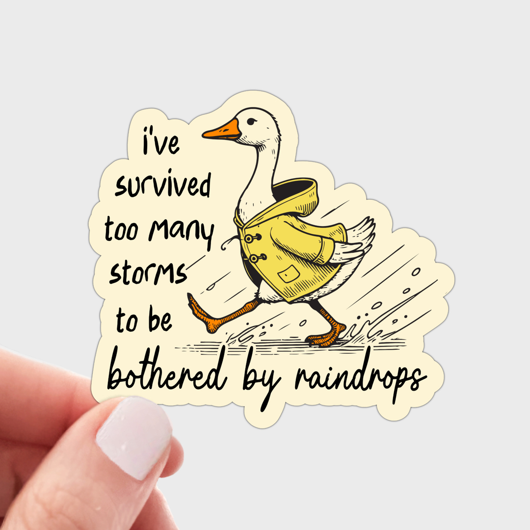 A die-cut sticker held by a hand, featuring an illustration of a white goose wearing a yellow raincoat walking through rain, next to the text 'i've survived too many storms to be bothered by raindrops'.