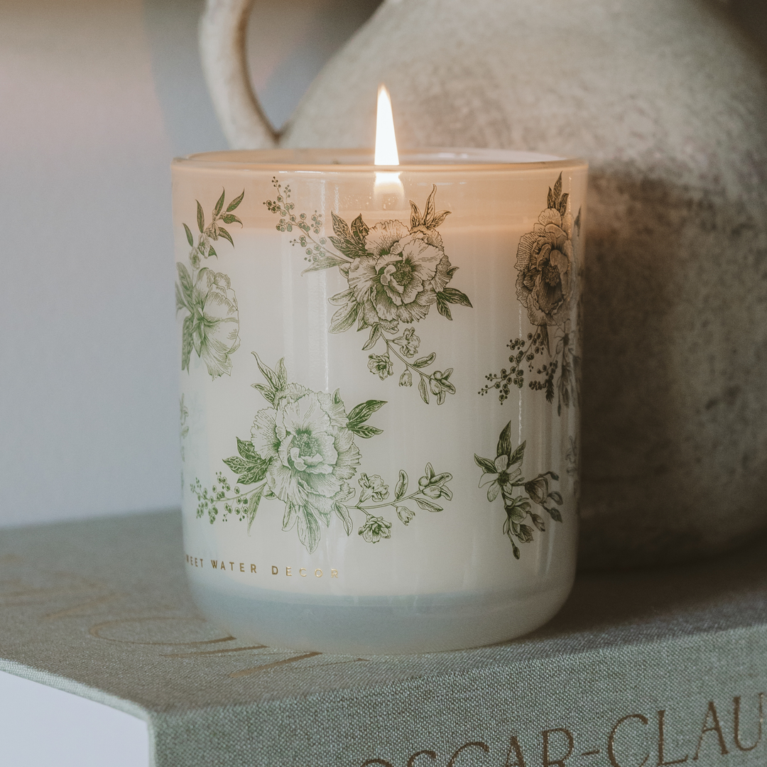 Lit candle with green delft design placed on a book and next to a pitcher. 
