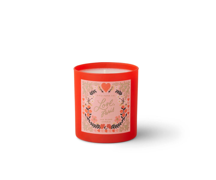 Candle in red glass container with label of a floral design with text 'Love, Paris' in gold script. 