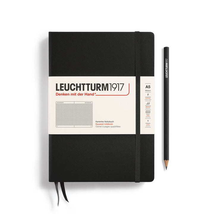 A Leuchtturm1917 A5 hardcover squared page notebook in black with a white label on the front, shown closed with an elastic closure and ribbon bookmark. A matching black pencil is placed vertically to the right.