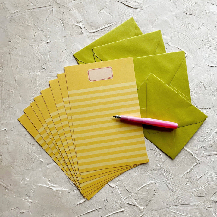 Eight sheets of yellow paper with horizontal stripes fanned on top of green envelopes. Each sheet has a yellos band at the top and the drawing of a label reminiscent of the cover of essay books.  