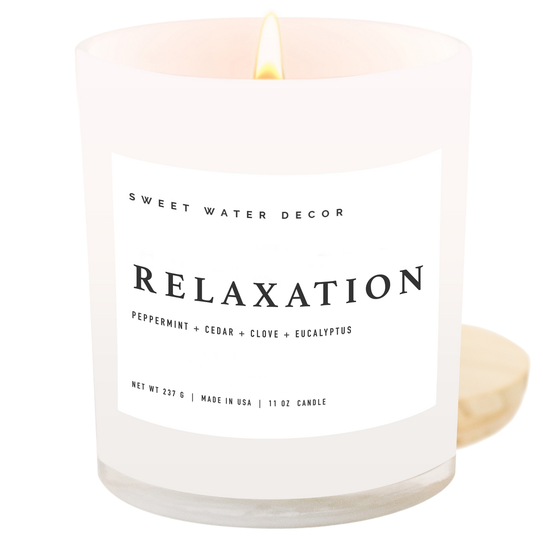 Lit candle in white opaque container with white label next to its wooden cap.