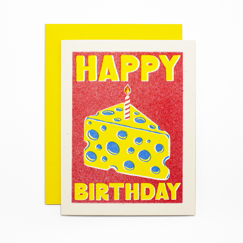 A birthday greeting card with a bright red glitter-textured background, yellow 'HAPPY BIRTHDAY' lettering, and a yellow slice of cheese with blue holes and a single candle. A matching yellow envelope is placed behind the card.