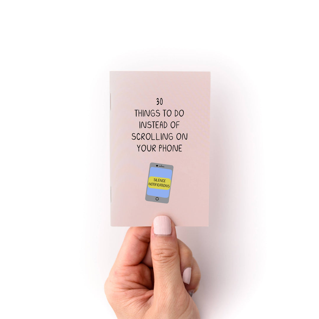 Hand holding a small paper book with text '30 Things to Do Instead of Scrolling on Your Phone' and a hand-drawn illustration of a mobile phone.