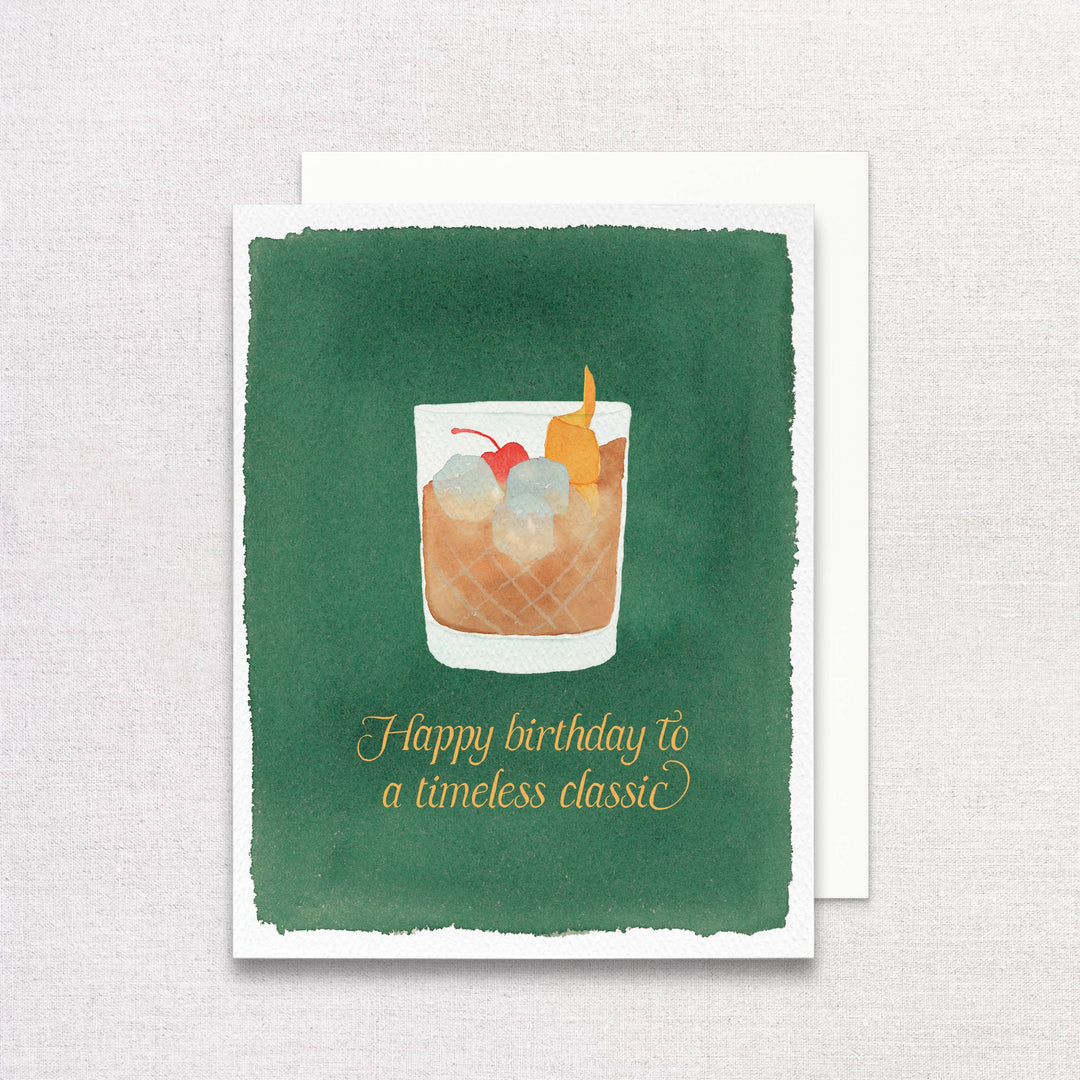 Front of card with a glass of an old fashioned cocktail, with cherry and orange twist, above gold script text on a green background with white envelope.