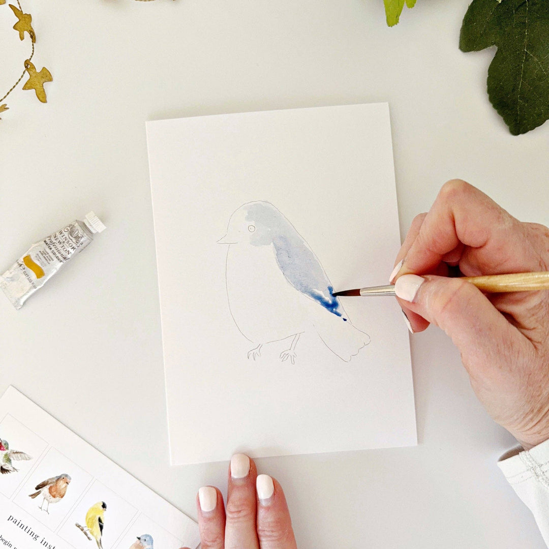 Person painting a bird outline with a paintbrush, surrounded by art supplies.