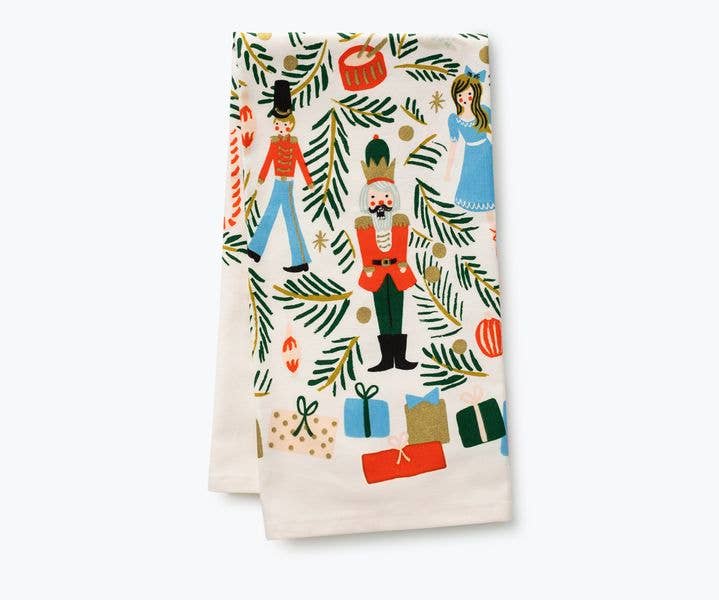 Folded tea towel with Nutcracker characters, green branches, and presents on an off-white background.