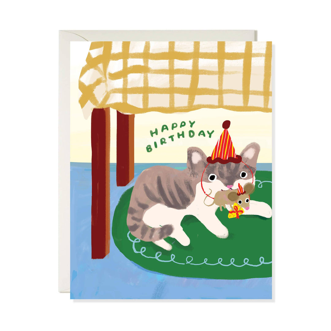 A birthday greeting card featuring an illustrated gray and white cat wearing a party hat and holding a small mouse under a yellow-and-white checkered tablecloth, with the words “Happy Birthday” on the card. A white envelope is visible behind the card.