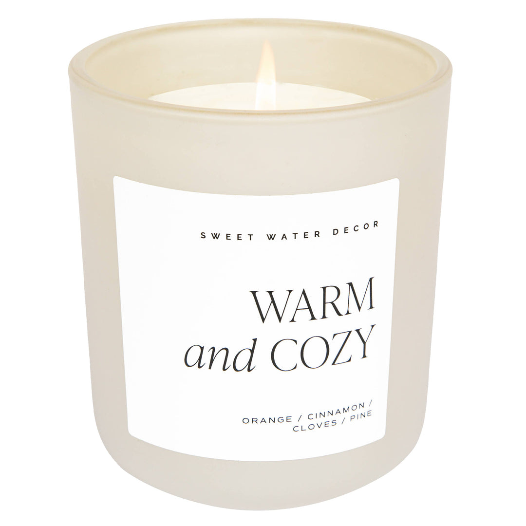 Scented soy candle in matte white glass container. Label is Warm and Cozy  Orange / Cinnamon / Cloves / Pine