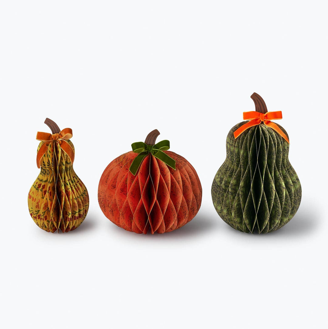 Three decorative honeycomb paper gourds arranged side by side. The left gourd is yellow and green striped with an orange bow, the middle pumpkin is orange with a green bow, and the right gourd is dark green with an orange bow.