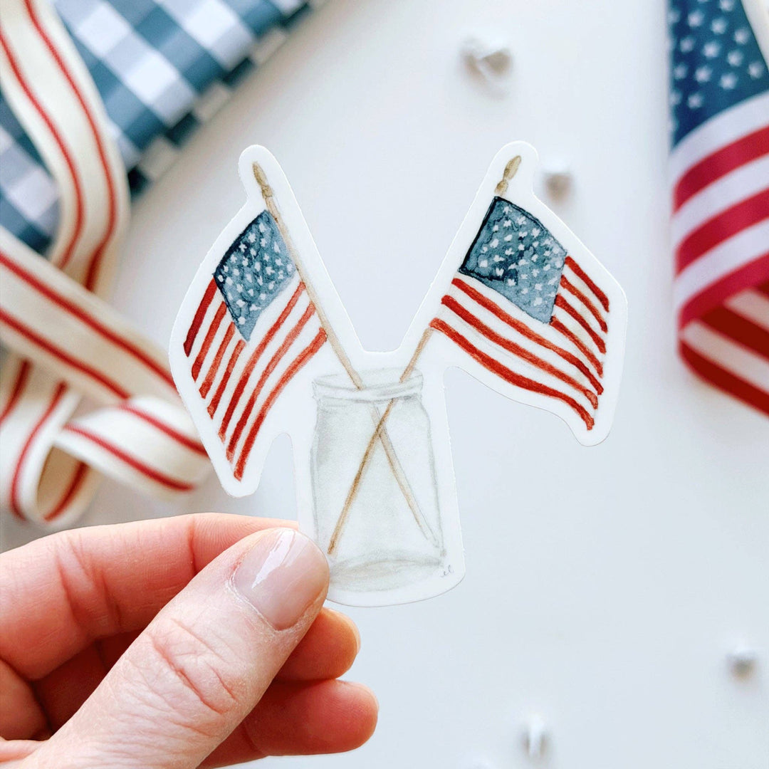 Hand holding a sticker of two American flags in a clear botte, on a background of patriotic objects.