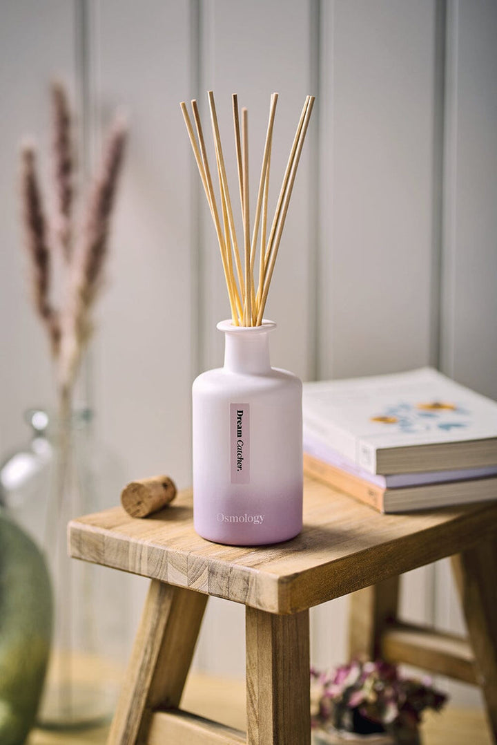 Diffuser container on a bench. 9 reeds are set in a round opaque narrow-neck bottle, in pink ombre gradient. 