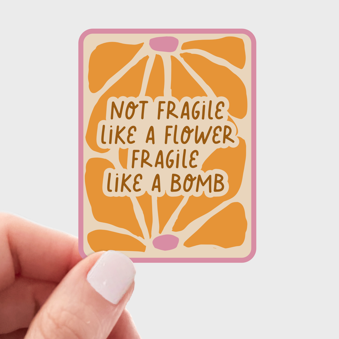 Hand holding a rectangular sticker with light yellow background and light pink border, with dark yellow flower buds above and below the text 'not fragile like a flower, fragile like a bomb.'