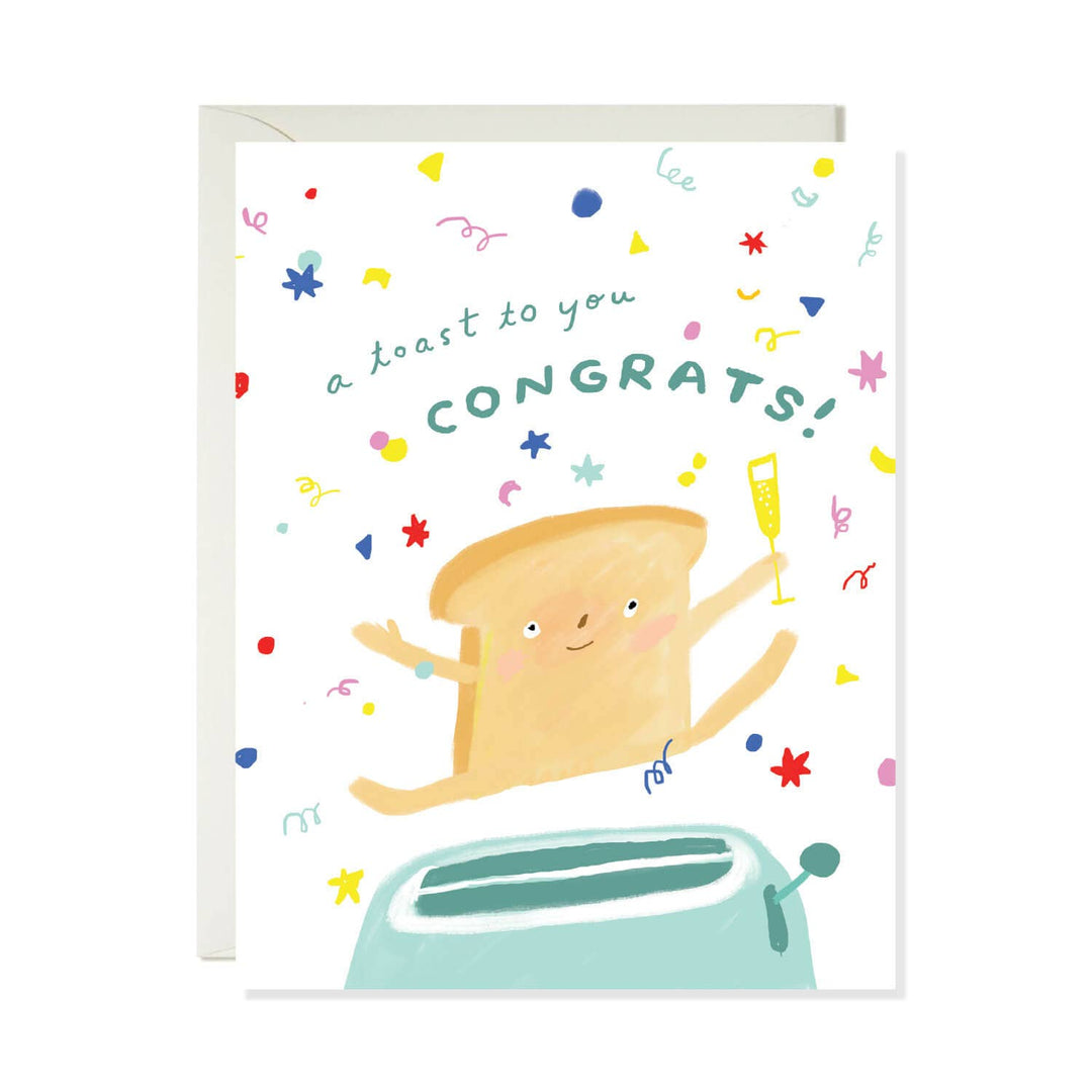 A white greeting card with a playful illustration of a slice of toast jumping from a mint-green toaster, surrounded by colorful confetti and the text 'a toast to you CONGRATS!'. A light gray envelope is visible behind the card.