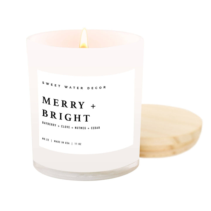 Scented soy candle in matte white glass container with wooden lid. Label is Merry + Bright  Bayberry / clove / nutmeg / cedar