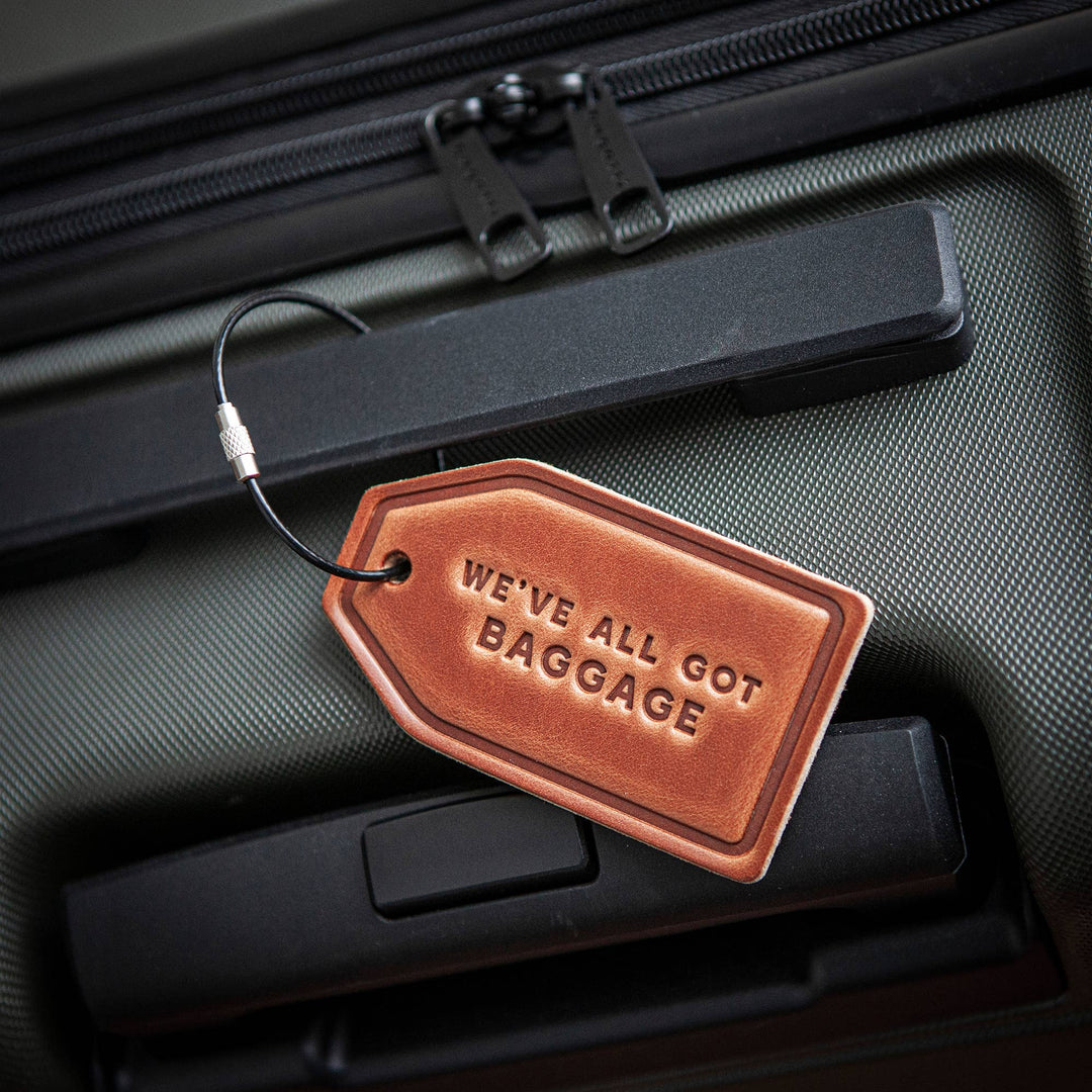 Brown leather luggage tag with 'WE'VE ALL GOT BAGGAGE' text on a suitcase.