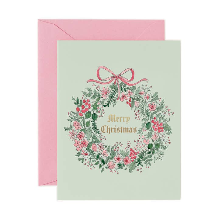 A square Christmas greeting card with a pale green background, featuring a watercolor-style floral wreath with pink flowers, green leaves, and a pink bow. The card reads 'Merry Christmas' in gold lettering and is shown with a pink envelope behind it.