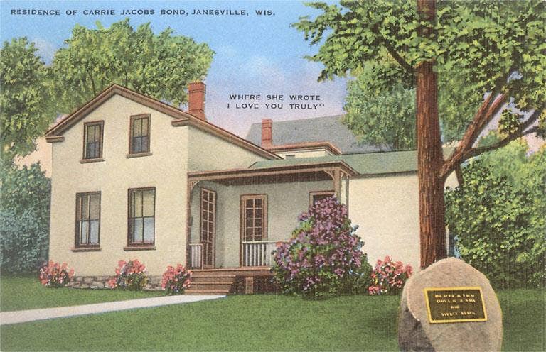 Pastel drawing of the Carrie Jacobs Bond house in Janesville, Wisconsin. It is a modest stucco two-story house with a lilac bush and other ornamental shrubs  around the front and a large boulder with a plaque in the foreground.