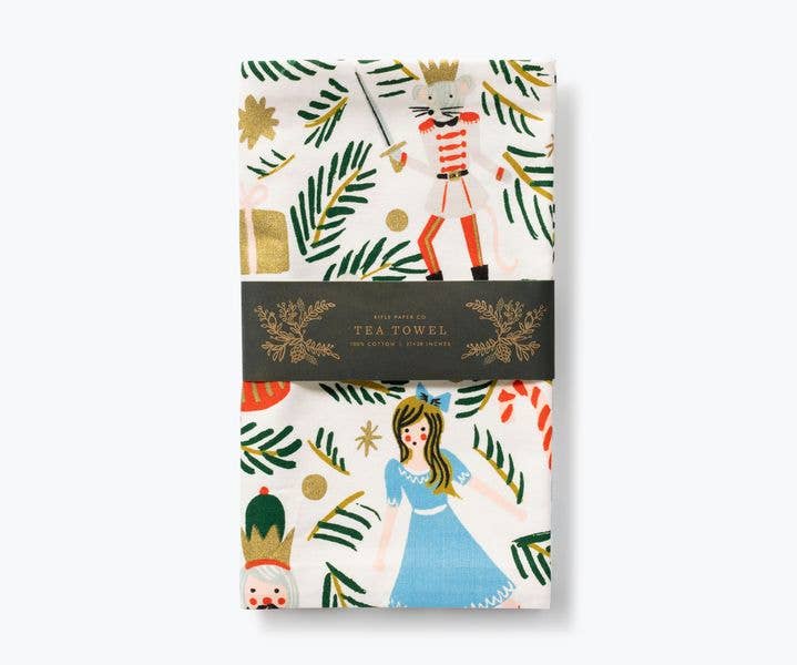 Folded tea towel with Nutcracker characters, green branches, and presents on an off-white background with paper packaging band.