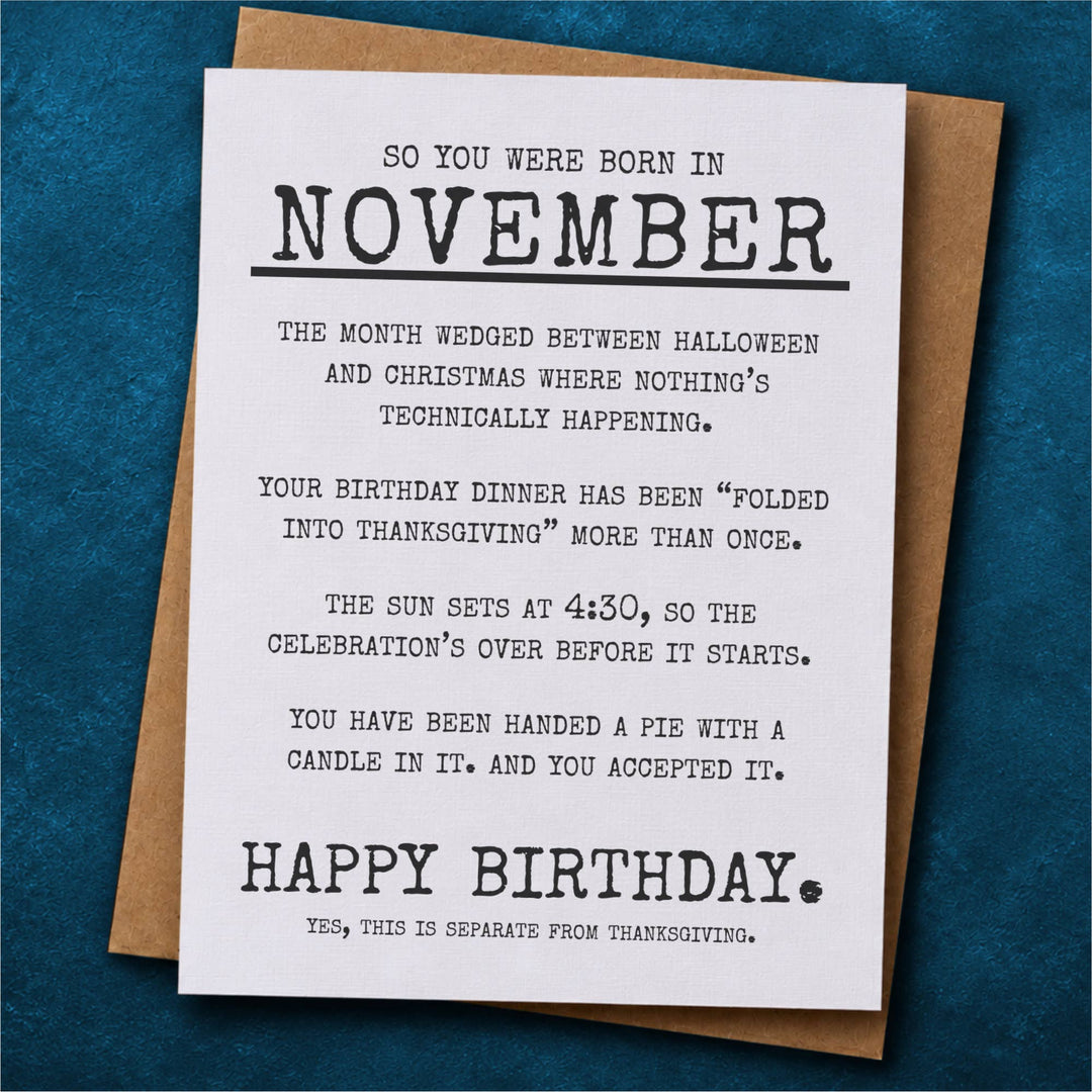 A novelty birthday card with a humorous Thanksgiving-themed message for November birthdays, displayed on top of a brown kraft envelope.