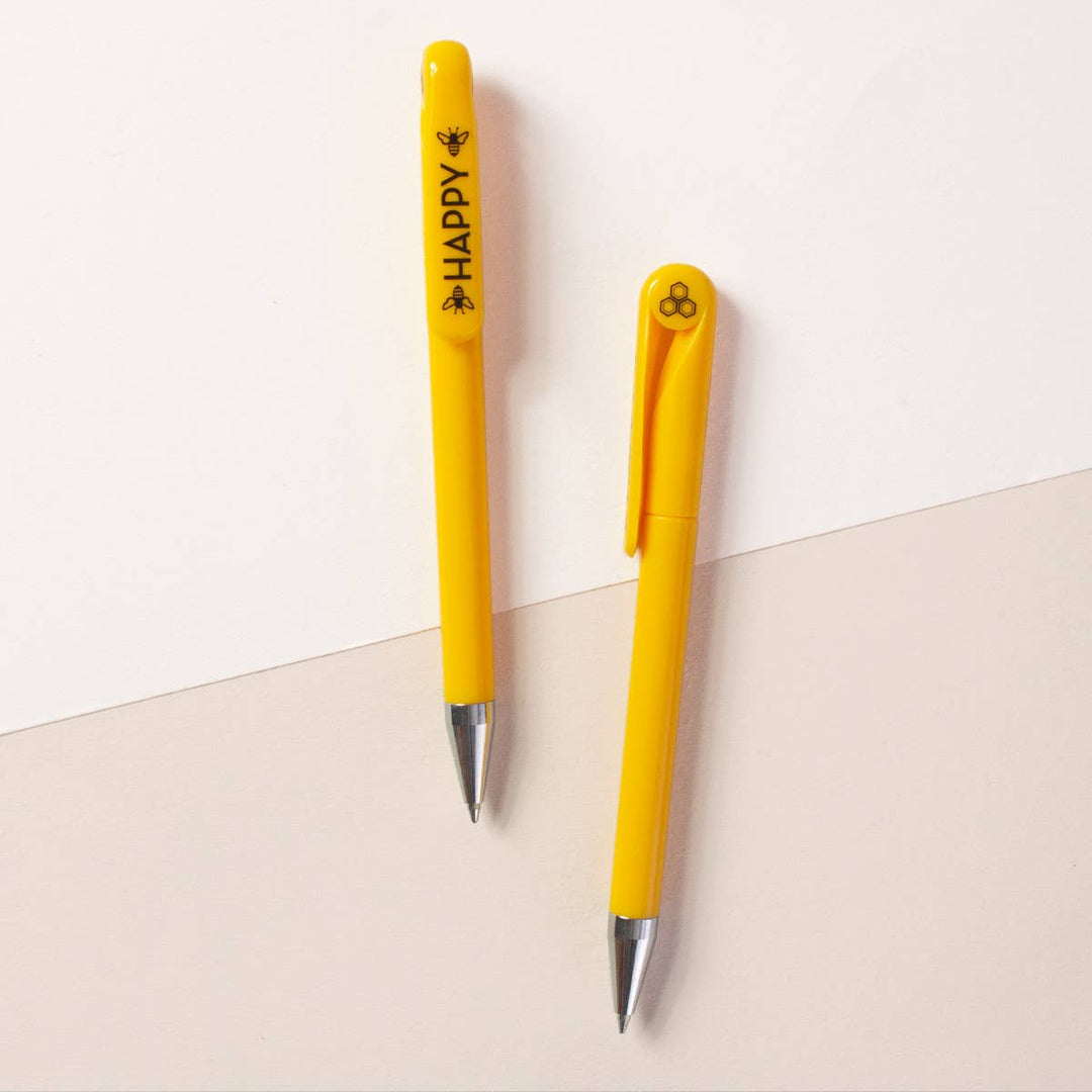 Two sides of a yellow retractable ballpoint pen with silver tips. One side shows the word 'Happy'' in black capital letters with a bee on either side, and the other side has a circular white cap insert with an image of a honeycomb.