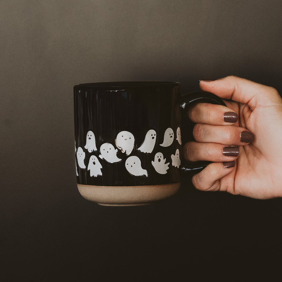 Female hand holding a black mug with two rows of variously shaped white ghost designs around the bottom.