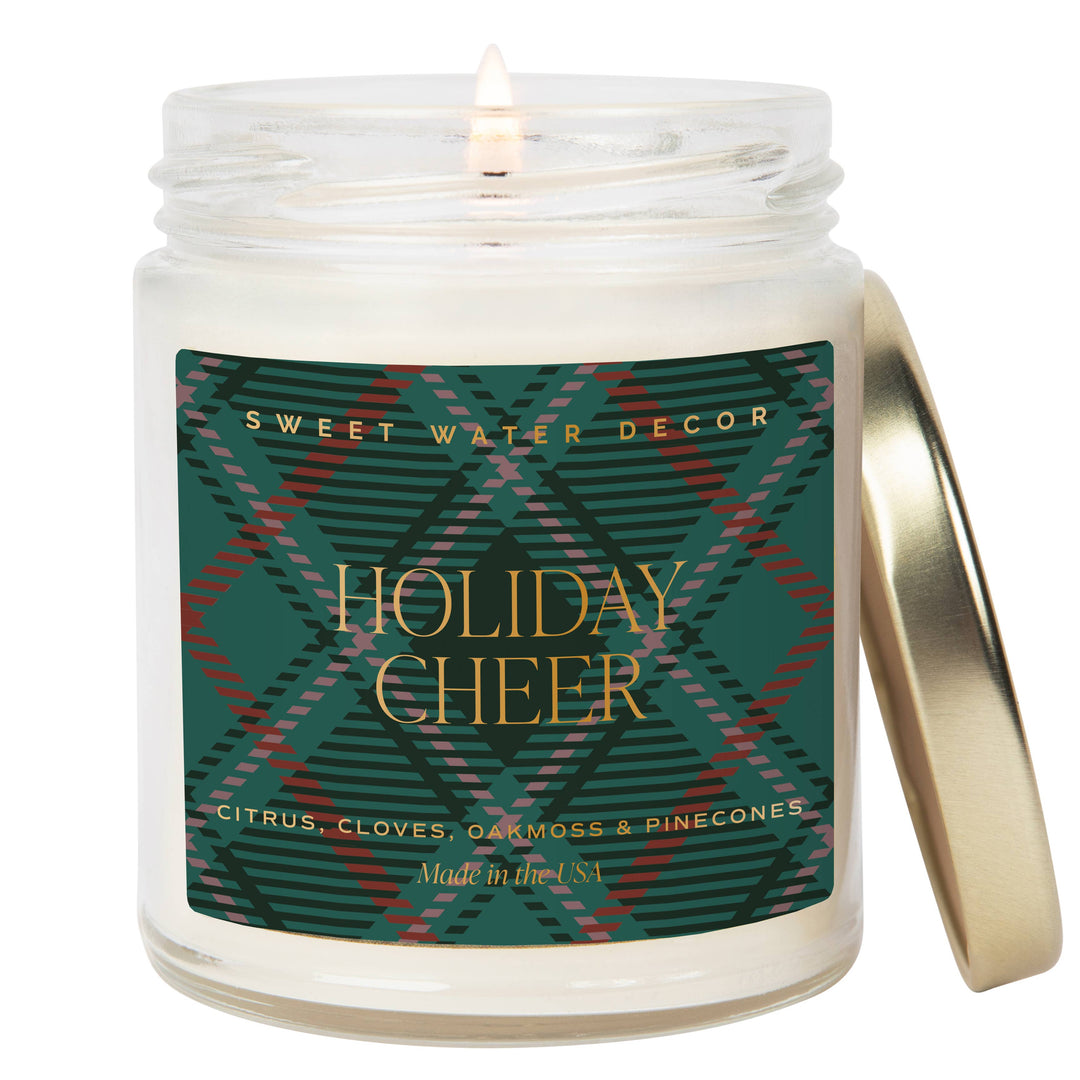 Scented soy candle in clear glass container with decorative label. Label is Holiday Cheer citrus / cloves / oak moss / pinecones