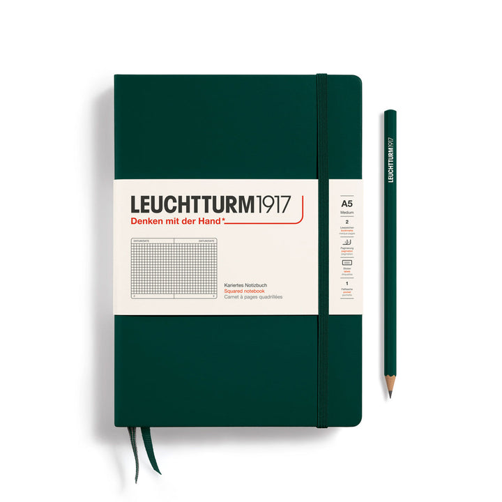 A Leuchtturm1917 A5 hardcover squared page notebook in forest green with a white label on the front, shown closed with an elastic closure and ribbon bookmark. A matching forest green pencil is placed vertically to the right.
