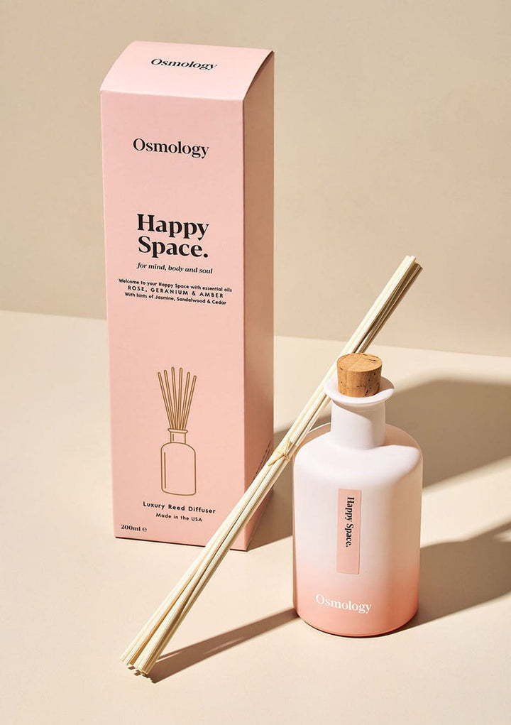 Diffuser container on a bench. 9 reeds lean against a round opaque narrow-neck bottle with cork stopper, in pink ombre gradient