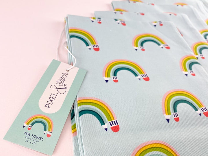 Close up of a colorful rainbow-patterned kitchen towel with pencils  arced to appear as a rainbow, on a light blue background.