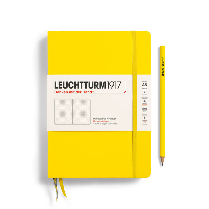 A Leuchtturm1917 A5 hardcover dotted page notebook in lemon yellow with a white label on the front, shown closed with an elastic closure and ribbon bookmark. A matching lemon yellow pencil is placed vertically to the right.