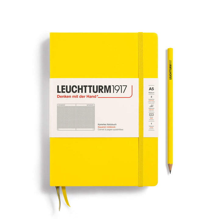 A Leuchtturm1917 A5 hardcover squared page notebook in lemon yellow with a white label on the front, shown closed with an elastic closure and ribbon bookmark. A matching lemon yellow pencil is placed vertically to the right.