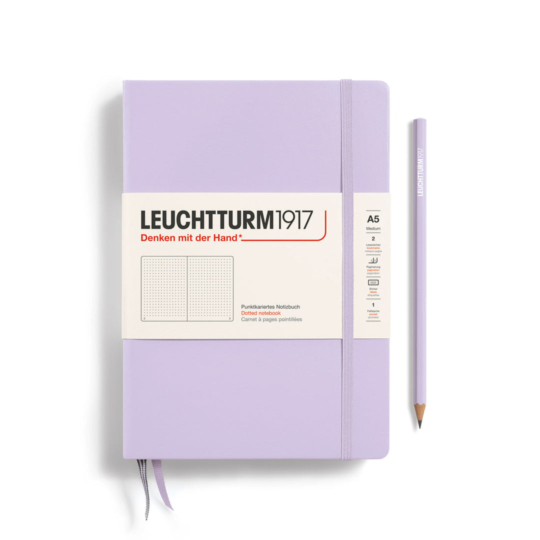 A Leuchtturm1917 A5 hardcover dotted page notebook in lilac with a white label on the front, shown closed with an elastic closure and ribbon bookmark. A matching lilac pencil is placed vertically to the right.