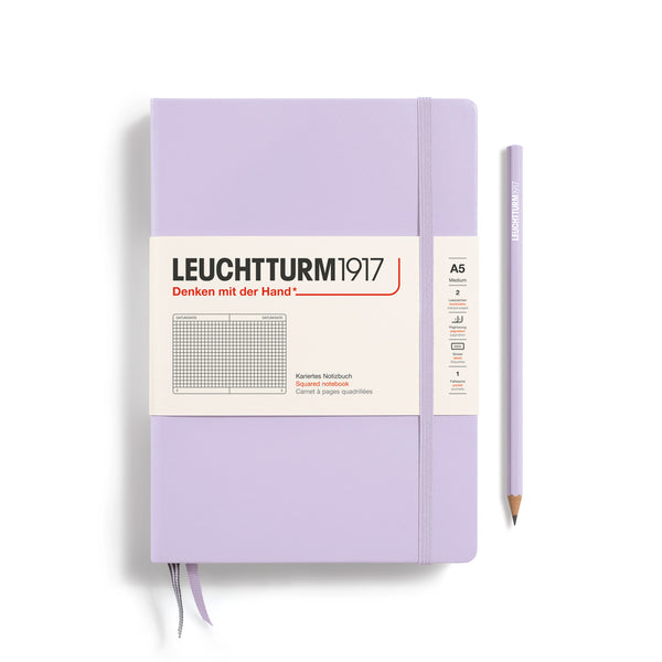 A Leuchtturm1917 A5 hardcover squared page notebook in lilac with a white label on the front, shown closed with an elastic closure and ribbon bookmark. A matching lilac pencil is placed vertically to the right.