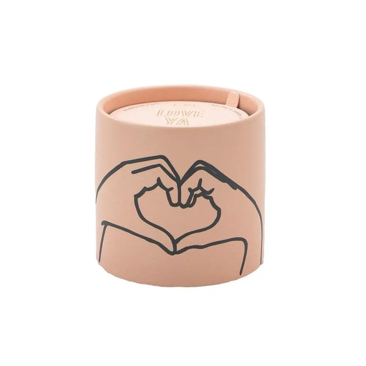 Scented candle in pinkish container with image of two hands making the heart symbol.