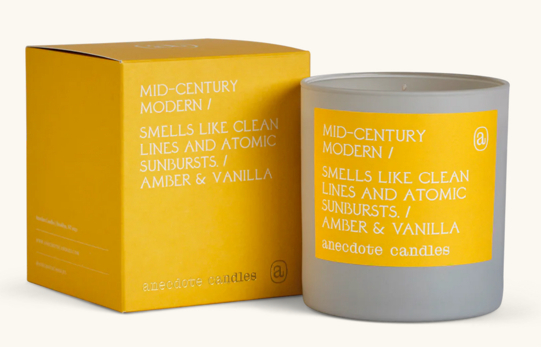 Scented candle in a stylish gray glass jar  next to its gift box. Yellow label with white text is Mid-Century Modern / Smells like Clean Lines and Atomic Sunbursts / Amber & Vanilla