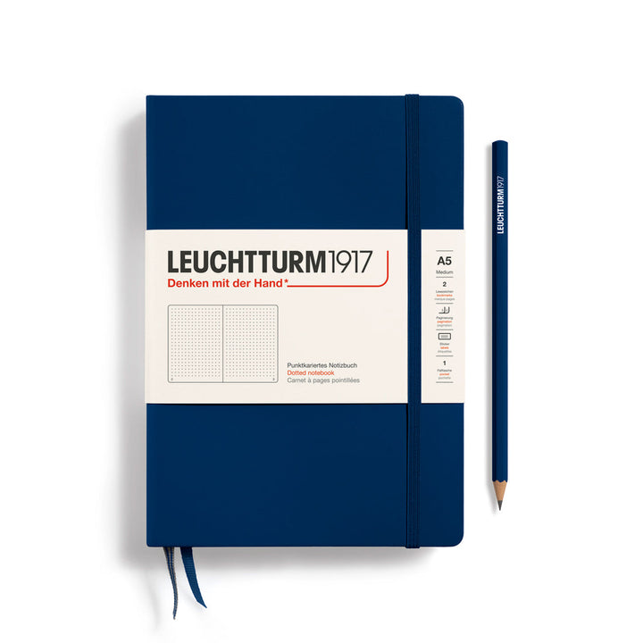 A Leuchtturm1917 A5 hardcover dotted page notebook in navy blue with a white label on the front, shown closed with an elastic closure and ribbon bookmark. A matching navy blue pencil is placed vertically to the right.