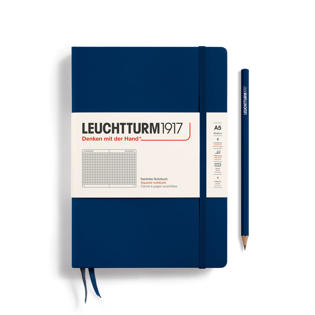 A Leuchtturm1917 A5 hardcover squared page notebook in navy blue with a white label on the front, shown closed with an elastic closure and ribbon bookmark. A matching navy blue pencil is placed vertically to the right.