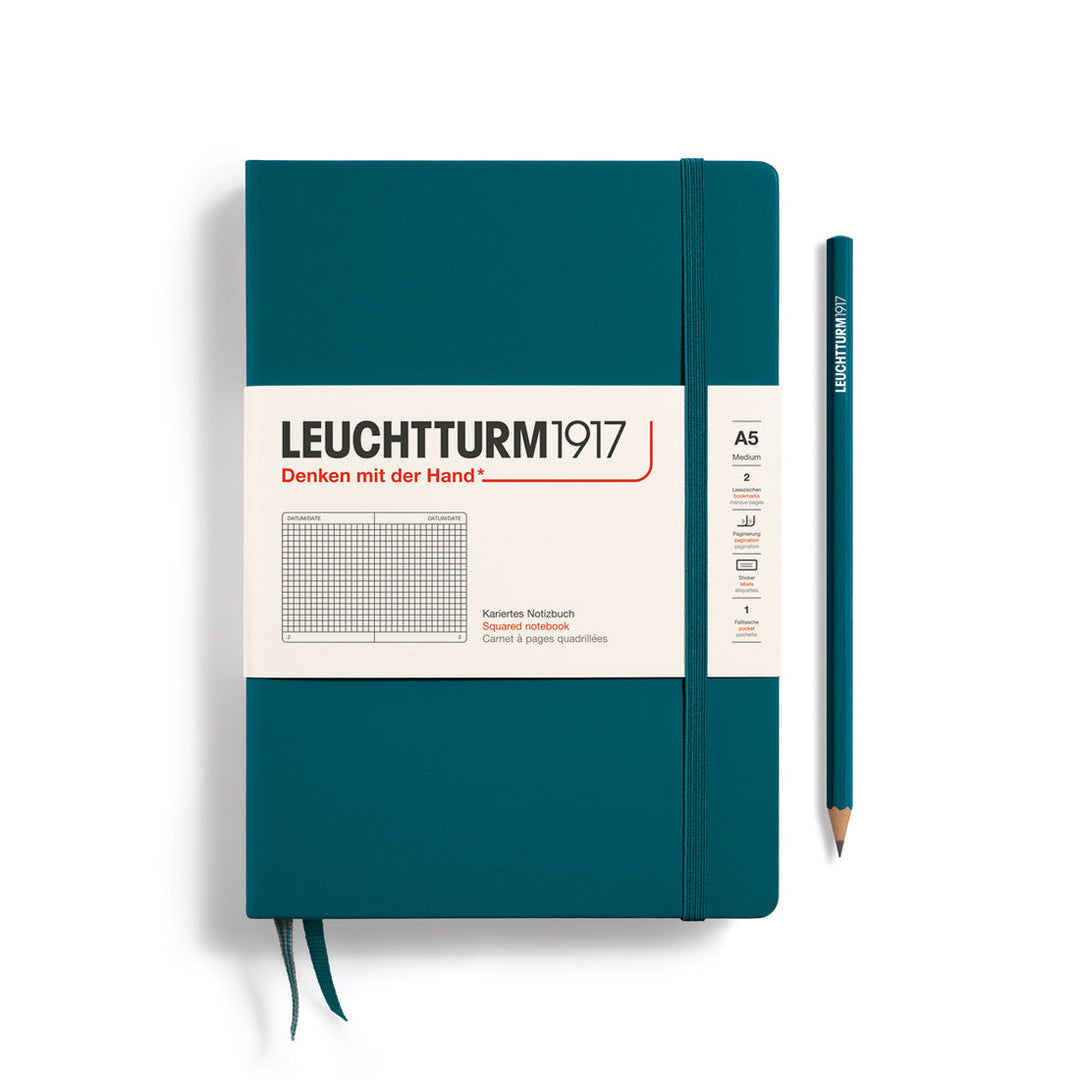 A Leuchtturm1917 A5 hardcover squared page notebook in pacific green with a white label on the front, shown closed with an elastic closure and ribbon bookmark. A matching pacific green pencil is placed vertically to the right.