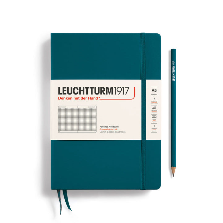 A Leuchtturm1917 A5 hardcover squared page notebook in pacific green with a white label on the front, shown closed with an elastic closure and ribbon bookmark. A matching pacific green pencil is placed vertically to the right.