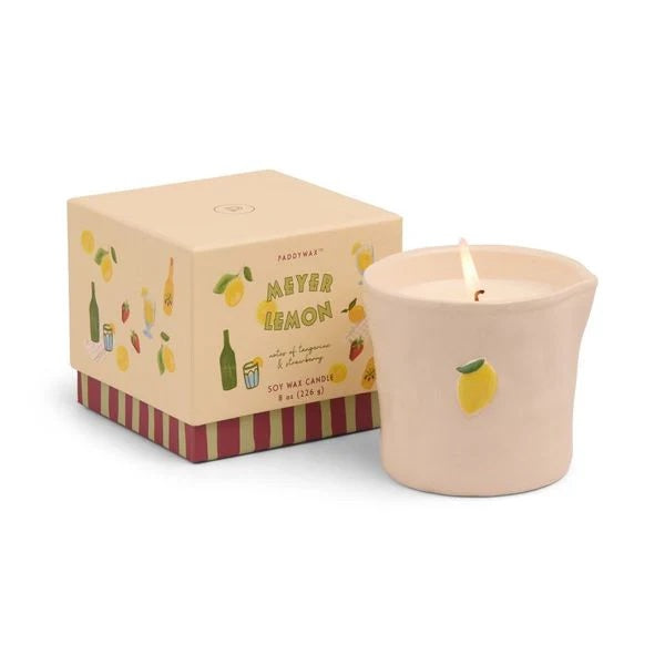 Burning candle in a beige ceramic vessel decorated with a single lemon, next to a gift box with images of lemons, sparkling water bottles, and strawberries.