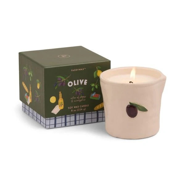 Burning candle in a beige ceramic vessel decorated what a single olive, next to a gift box with images of olives, figs, vinegar bottles and baguettes.