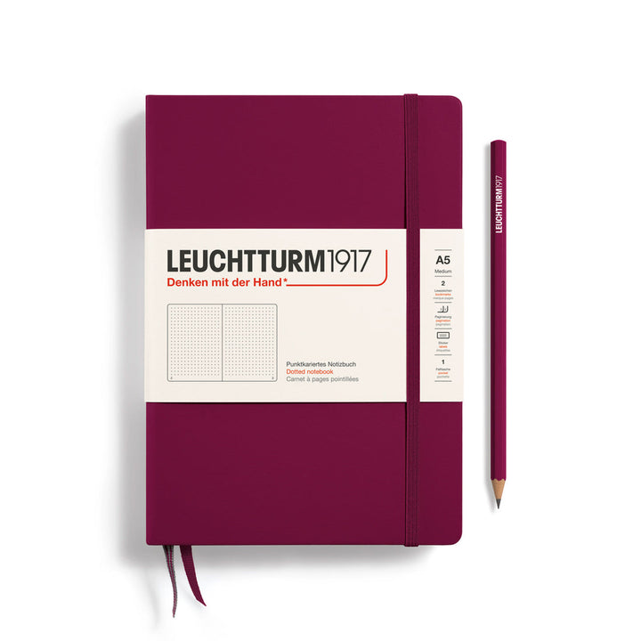A Leuchtturm1917 A5 hardcover dotted page notebook in port red with a white label on the front, shown closed with an elastic closure and ribbon bookmark. A matching port red pencil is placed vertically to the right.
