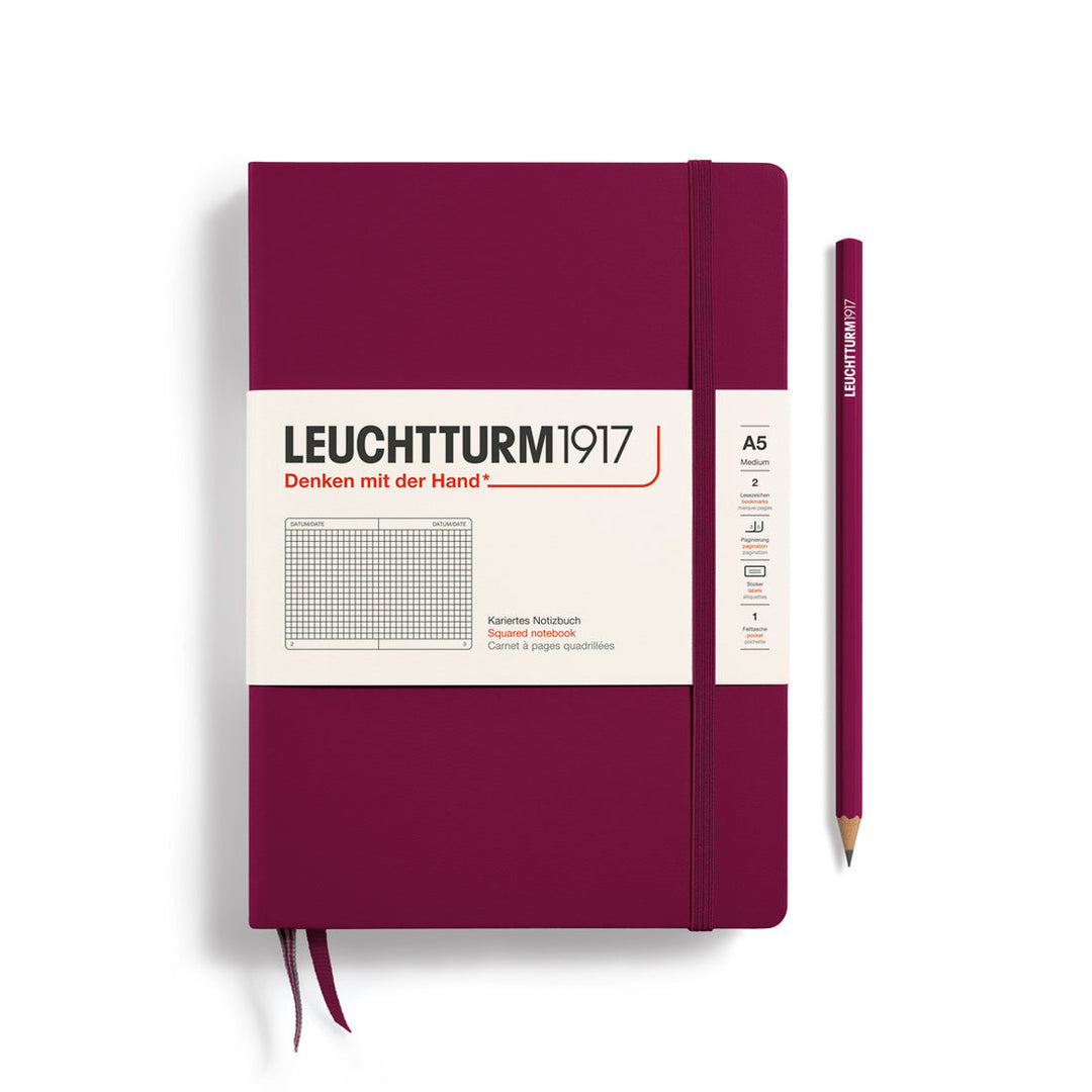 A Leuchtturm1917 A5 hardcover squared page notebook in port red with a white label on the front, shown closed with an elastic closure and ribbon bookmark. A matching port red pencil is placed vertically to the right.