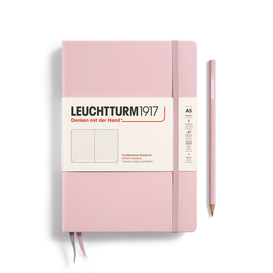 A Leuchtturm1917 A5 hardcover dotted page notebook in powder pink with a white label on the front, shown closed with an elastic closure and ribbon bookmark. A matching powder pink pencil is placed vertically to the right.