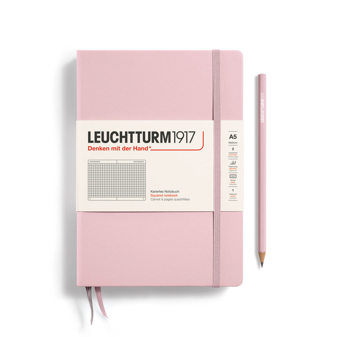 A Leuchtturm1917 A5 hardcover squared page notebook in powder pink with a white label on the front, shown closed with an elastic closure and ribbon bookmark. A matching powder pink pencil is placed vertically to the right.