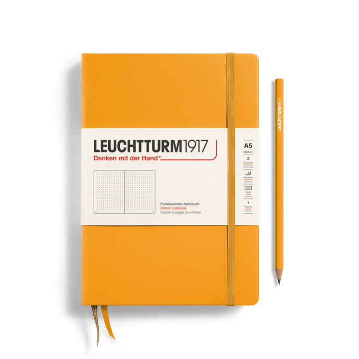 A Leuchtturm1917 A5 hardcover dotted page notebook in rising sun orange with a white label on the front, shown closed with an elastic closure and ribbon bookmark. A matching rising sun orange pencil is placed vertically to the right.