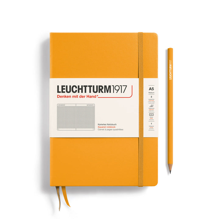 A Leuchtturm1917 A5 hardcover squared page notebook in rising sun orange with a white label on the front, shown closed with an elastic closure and ribbon bookmark. A matching rising sun orange pencil is placed vertically to the right.